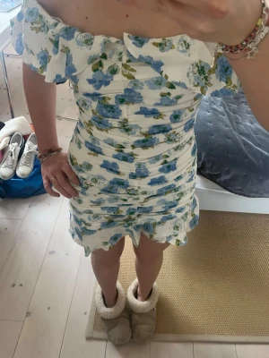 Blommig offshoulder kortklänning - Superfin kort klänning med offshoulder och volangdetaljer. Vit botten med blåa och gröna blommor, figurnära passform och rynkade sömmar som ger en snygg siluett. Perfekt för sommarens alla tillfällen!