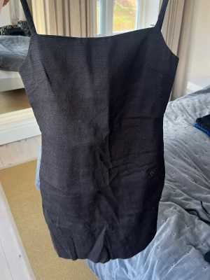 Svart kort klänning från Zara - Säljer en svart, stilren kort klänning från Zara i storlek XS. Klänningen har smala axelband, rak halsringning och en diskret dragkedja bak. Materialet är grovvävt och känns lite som linne, vilket ger en cool och enkel vibe.