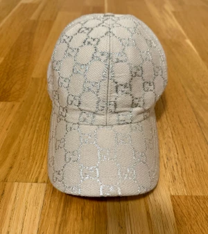 Beige och silver Gucci keps - Snygg beige keps från Gucci med glittrigt silvermönster i klassiskt GG-monogram. Utsidan är i grovvävd bomull och insidan har lyxig ljusblå satin. Justerbar baktill med svart band och metallring. Perfekt för dig som vill sticka ut med designerstil.