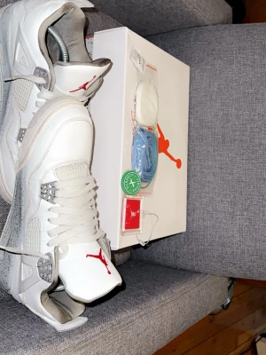 Nike Air Jordan 4 White Cement sneakers - Nike Air Jordan 4 White Cement sneakers i vit och grå med röd Jumpman-logga på plösen. Skorna har snörning, platt sula och rund tå. Ovandelen är i skinn med meshpaneler och detaljer i plast. Sulan har grått stänkmönster och synlig Air-enhet. Extra skosnören medföljer.