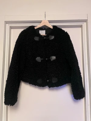 Svart teddyjacka med knappar - Mysig och skön svart teddyjacka med fluffig struktur och snygga dekorativa knappar framtill. Jackan är croppad och har långa ärmar. Materialet är teddy/fleece och detaljerna är i svart skinnimitation. Mycket bra skick, som ny! Sälj på flera sidor, först till kvarn. 