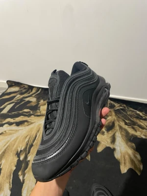 Nike Air Max 97 Triple Black sneakers - Nike Air Max 97 Triple Black sneakers med vågiga linjer och synlig Air-enhet i sulan. Hela skon är svart med mesh och syntetmaterial, samt diskret Nike-logga på sidan och tungan. Platt sula med mönstrad undersida för extra grepp. Snygg och sportig design.