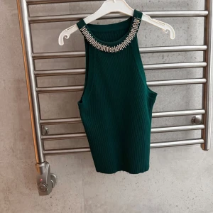 Grön ribbad linnetopp med pärlor - Snygg mörkgrön ribbad linnetopp med ärmlös design och rund halsringning dekorerad med glittrande pärlor. Perfekt att styla till jeans eller kjol för en cool look. Materialet är mjukt och stretchigt för skön passform.