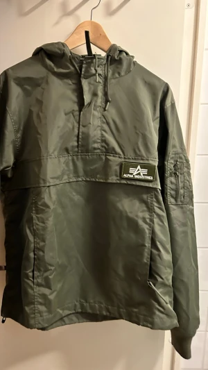 Alpha Industries Jacks  - Säljer en grön vindjacka från Alpha Industries med huva och snörning. Jackan har en halvlång dragkedja framtill, praktisk ficka på ärmen och tydlig logotyp-patch på bröstet. Materialet är slitstark polyester och passformen är normal.       Använd max 2-3 gånger så är i nyskick. 