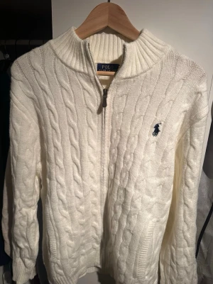 Vit stickad tröja från Polo Ralph Lauren (nypris över 3000kr) - Vit kabelstickad tröja från Polo Ralph Lauren med half zip och broderad logga på bröstet. Tröjan har en hög krage och långa ärmar, perfekt för kyliga dagar. Klassisk och stilren design som funkar till det mesta.