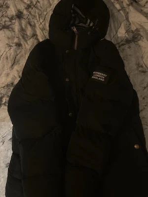 Svart pufferjacka från Burberry (ÄKTA) - Säljer en svart pufferjacka från Burberry med huva och tydlig logga på ärmen. Jackan har dragkedja och knappar framtill samt en klassisk, oversized look. Perfekt för kalla dagar och riktigt snygg till streetwear-stilen.