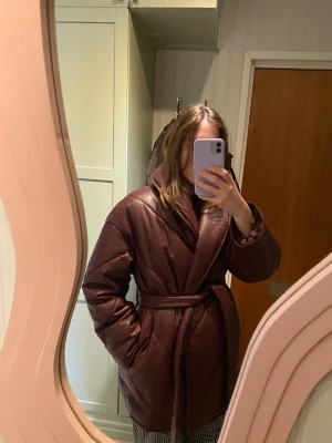 Quiltad vinröd pufferjacka från Monki - Säljer en vinröd quiltad pufferjacka från Monki med bälte i midjan. Jackan har oversized passform, bred krage och knäppning framtill. Materialet är syntet med skinnlook och den är perfekt för kalla dagar. Riktigt snygg och trendig jacka som sticker ut!