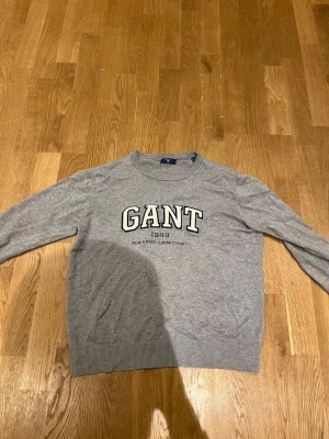 Grå stickad tröja från GANT - Snygg grå stickad tröja från GANT med klassisk rund hals och broderad logga framtill. Tröjan har långa ärmar och ribbade muddar vid hals, ärmslut och nederkant. Perfekt för dig som gillar stilren och enkel design med en sportig touch. Storlek 158/164. Pris kan diskuteras.