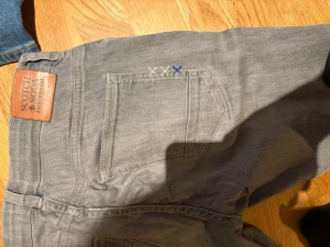 Grå jeans från Scotch & Soda - Säljer ett par grå jeans från Scotch & Soda Amsterdam med snygga broderade detaljer i blått och gult ovanför bakfickan. Klassisk femficksmodell med raka ben och normal passform. Materialet är mjukt jeans och färgen är ljusgrå.
