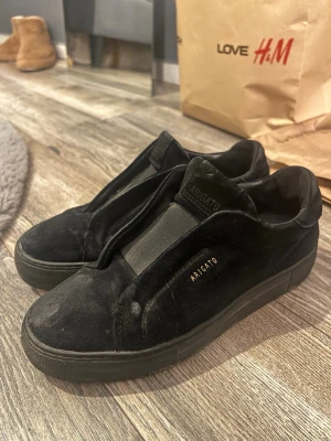Svarta sneakers från Axel Arigato - Säljer ett par svarta sneakers från Axel Arigato med stilren design och diskret logga i guld på sidan. Skorna har en rund tå, platt sula och är tillverkade i mocka med elastisk panel över vristen. Perfekta för dig som gillar minimalistisk stil. Fläckarna går att skrubba bort och denna modell säljs ej längre därav pris
