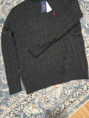   tröja Polo Ralph Lauren - Svart kabelstickad tröja från Polo Ralph Lauren med rund hals och röd broderad logga på bröstet. Tröjan har ribbade muddar vid ärmslut och nederkant, långärmad och klassisk passform. Perfekt för dig som gillar stilrena och tidlösa plagg.