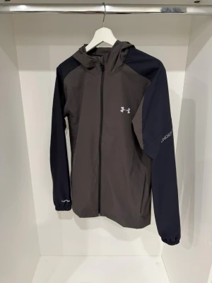 Under Armour grå och svart vindjacka - Snygg vindjacka från Under Armour i grått med svarta ärmar och huva. Jackan har dragkedja framtill, elastiska muddar och logga på bröstet samt ärmen. Perfekt för dig som gillar sportig stil och vill ha något lätt och funktionellt.