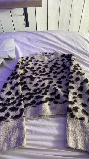 Beige leopardmönstrad stickad tröja - Säljer en beige stickad tröja med svart leopardmönster. Tröjan har breda ribbstickade muddar vid ärmslut och nederkant samt rund halsringning. Materialet ser mjukt och fluffigt ut, perfekt för kyligare dagar. Oversized passform för en avslappnad look.