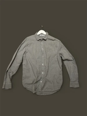 Grå långärmad skjorta i bomull - Stilren grå skjorta med klassisk krage och knäppning framtill. Skjortan är tillverkad i mjuk bomull och har långa ärmar med manschetter. Perfekt för en clean och enkel look.