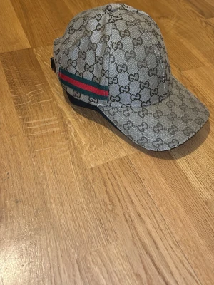 Gucci beige keps med GG-mönster - Snygg beige keps från Gucci med klassiskt GG-monogram över hela kepsen och en ikonisk grön-röd rand på sidan. Kepsen har böjd skärm och justerbar rem bak. Tillverkad i canvasmaterial som ger en exklusiv känsla.