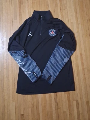 Svart PSG träningströja Jordan - Svart långärmad träningströja från Jordan x PSG med halv dragkedja, PSG-logga på bröstet och vita detaljer. Ärmarna har grått grafiskt mönster och tumhål. Perfekt för fotbollsträning eller sportiga dagar. Egentligen är storleken L men det passar också för S och M