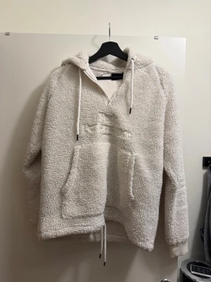 Vit teddyhoodie från Peak Performance - Mysig vit teddyhoodie från Peak Performance med halv dragkedja, broderad logga på bröstet och stor känguruficka. Hoodien har dragsko i huvan och nederkanten samt ribbade muddar vid ärmsluten. Perfekt för dig som gillar en avslappnad och trendig stil.