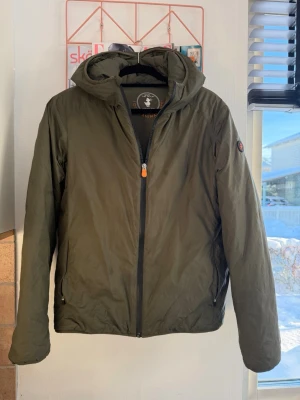 Olivgrön vindjacka från Save The Duck - Snygg olivgrön vindjacka från Save The Duck med huva och hel dragkedja framtill. Jackan är lättviktig och har två fickor med dragkedja. Tillverkad i syntetmaterial och är helt fri från animaliska produkter. Se fläck på bild. Inget som stör. 