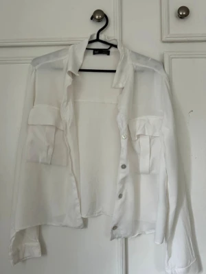 Vit kofta med fickor från Zara  - Stilren vit skjorta från zara med två stora bröstfickor och knäppning framtill. Skjortan har lång ärm och är tillverkad i ett tunt, lätt material som ger en luftig känsla. Perfekt för en avslappnad och trendig look.