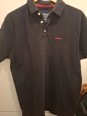 Svart pikétröja från Sebago - Klassisk svart pikétröja från Sebago Marine Classics med röd logga broderad på bröstet. Tröjan har korta ärmar, krage och knappar framtill samt diskreta röda detaljer på insidan av kragen. Perfekt för en clean och stilren look. Den är lite blekt pågrund av att den tvättad några gånger. Priset går att diskutera skrev om du har frågor 