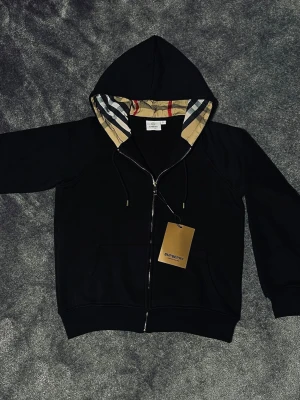 Svart Burberry zip hoodie - Säljer en svart hoodie från Burberry. Huvan har det klassiska Burberry-rutiga mönstret på insidan. Materialet är mjukt och bekvämt, perfekt för en snygg och avslappnad stil. Tröjan är helt ny o aldrig använd. Det är bara att skriva ifall du har fler frågor!