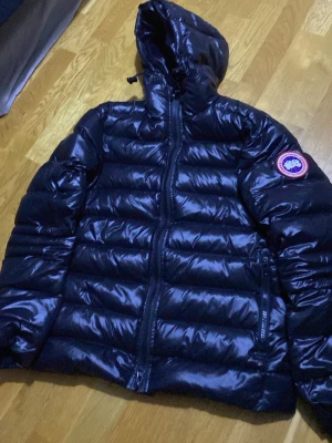 Svart dunjacka från Canada Goose - Snygg svart dunjacka från Canada Goose med glansig finish, huva och dragkedja framtill. Jackan har märkets klassiska logga på ärmen och invändiga axelband med Canada Goose-tryck. Perfekt för kalla dagar.