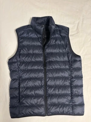 Uniqlo dunväst - Snygg mörkblå dunväst med hög krage och horisontella sömmar. Västen har en stilren design med dragkedja framtill och två sidofickor. Perfekt att slänga över en hoodie eller tröja för extra värme. Materialet är glansigt och lätt, vilket gör den enkel att bära.