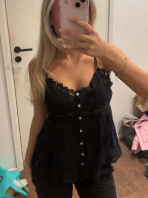 Svart linnetopp med spets och knappar - Svart linnetopp med tunna axelband, spetsdetaljer vid urringningen och knäppning framtill. Luftig och flowy passform, perfekt för en trendig look.