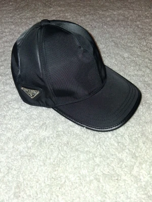 Svart keps från Prada i nylon - Snygg svart keps från Prada i glansigt nylonmaterial med klassisk böjd skärm och ikonisk metallogo på sidan. Perfekt accessoar för en trendig streetstyle-look.