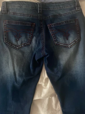 Mörkblå jeans med broderade fickor - Snygga mörkblå jeans från Gina tricot med coola röda broderier på bakfickorna och slitningar framtill. Klassisk femficksmodell med silverfärgade knappar och detaljer. Perfekta för dig som gillar streetstyle och vill sticka ut lite extra. Lågmidjade 