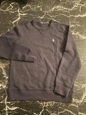 Mörkblå tröja från Polo Ralph Lauren - Snygg mörkblå långärmad tröja från Polo Ralph Lauren med klassisk rund halsringning och broderad vit logga på bröstet. Tillverkad i mjuk bomull som känns skön mot huden. Perfekt för dig som gillar stilren och enkel design.