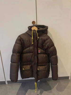 Brun dunjacka från Fjällräven - Mörkbrun dunjacka från Fjällräven med huva och gula dragskor. Jackan har två stora fickor framtill med lock och tryckknappar samt klassisk Fjällräven-logga på bröstet. Perfekt för kalla vinterdagar.