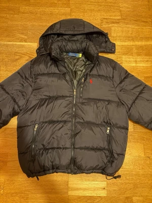 Svart Ralph Lauren dunjacka - Svart Polo Ralph Lauren dunjacka med röd logga i storlek XL.   Jackan är väldigt fin och i gott skick, finns en liten fläck på baksidan, har inte testat ta bort den. Midjan är justerbar.  Kontakta gärna för frågor och pris!💯