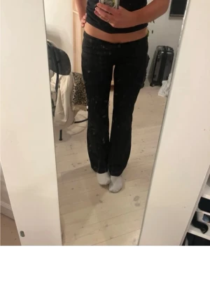 Svarta bootcut byxor - Säljer ett par svarta byxor med bootcut-modell och låg midja. Materialet är glansigt, byxorna har knapp och dragkedja framtill samt klassiska fickor.