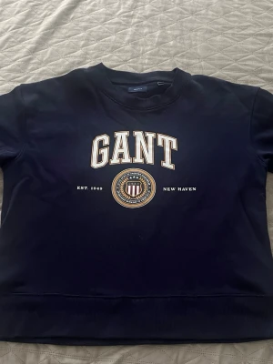 Mörkblå sweatshirt från GANT - Snygg mörkblå sweatshirt från GANT med klassiskt tryck framtill och rund hals. Tröjan har ribbade muddar vid ärmslut och nederkant samt ett emblem med texten 'EST. 1949' och 'NEW HAVEN'. Perfekt för en avslappnad och stilren look.