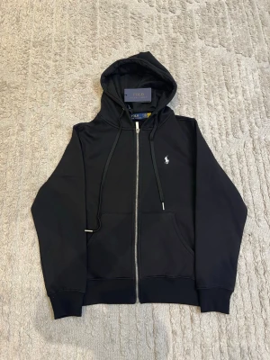 Svart hoodie från Polo Ralph Lauren - Svart hoodie med dragkedja från Polo Ralph Lauren. Klassisk design med huva och justerbara snören, två fickor framtill och den ikoniska vita logon broderad på bröstet. Ribbstickade muddar vid ärmslut och nederkant. Perfekt för en avslappnad stil.