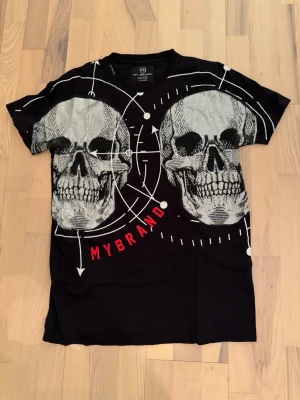 Svart t-shirt med döskallar från MyBrand - Cool svart t-shirt från MyBrand med två stora vita döskallar och grafiska detaljer framtill. Trycket har även röda bokstäver som stavar 'MYBRAND'. Klassisk rund hals och korta ärmar. Perfekt för dig som gillar edgy streetwear. Den är i bra skick endast att ”stenarna” har fallit av under armhålorna men inget som syns när man använder den. Storlek XL men passar mig som har L. Hör av dig vid fler frågor och funderingar, pris kan diskuteras 