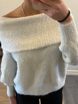 Vit off shoulder stickad tröja - Mysig vit stickad tröja med off shoulder-design och bred krage. Tröjan har en fluffig, mjuk struktur och långa ärmar med ribbade muddar. Perfekt för att skapa en avslappnad men trendig look.