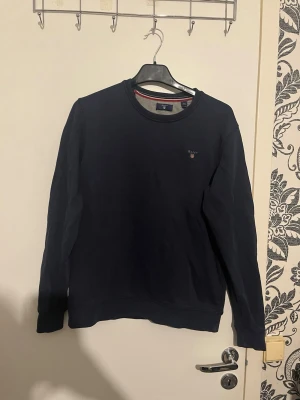 Mörkblå sweatshirt från Gant - Säljer en stilren mörkblå sweatshirt från Gant med rund halsringning och diskret logga broderad på bröstet. Tröjan har ribbade muddar vid ärmslut och nederkant samt en klassisk passform. Perfekt för en avslappnad och snygg look. OBS KÖPT FRÅN ZALANDO storlek xs (170) barnstorlek