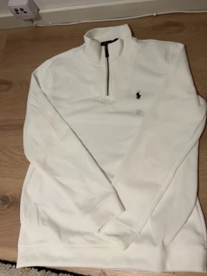 Vit quarter zip från Ralph Lauren - Snygg vit långärmad tröja från Ralph Lauren med quarter zip och hög krage. Tröjan har en broderad marinblå logga på bröstet och är tillverkad i mjukt material. Perfekt för en clean och stilren look.