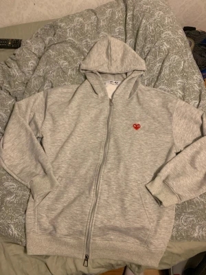 Grå hoodie från Comme des Garçons Play - Snygg grå zip-hoodie från Comme des Garçons Play med det ikoniska röda hjärtat med ögon broderat på bröstet. Hoodien har dragkedja, huva och ribbade muddar. Perfekt för en avslappnad och trendig stil.
