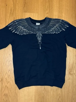 Svart sweatshirt Marcelo Burlon - Svart sweatshirt från Marcelo Burlon County of Milan med ikoniskt vingtryck i grått och vitt över axlar och halsringning. Rund hals och klassisk passform, perfekt för en edgy streetstyle-look.