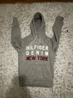 Grå Hilfiger Denim hoodie - Säljer en grå hoodie från Hilfiger Denim med stor vit och röd text på bröstet. Hoodien har huva med vita dragsnören. Storlek S och mycket bra skick, hör gärna av dig vid minsta fundering!