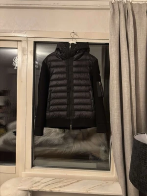 Moncler cardigan  - Snygg svart dunjacka med quiltad framsida och stickade ärmar. Jackan har en vadderad huva, dragkedja framtill och ribbade muddar. Perfekt för kalla vinterdagar och har en modern, sportig look. Använd, men inga defekter 