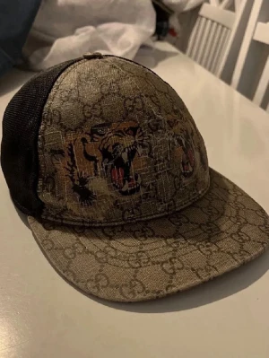 Gucci keps med tigertryck - Snygg keps från Gucci med klassiskt GG-mönster och coolt tigertryck framtill. Kepsen har svart mesh på baksidan och böjd skärm. Perfekt accessoar för att lyfta din streetwear-look.
