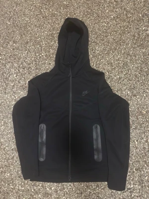 Svart Nike hoodie med dragkedja - Svart hoodie från Nike med hel dragkedja framtill och två fickor med dragkedja. Klassisk huva och diskret Nike-logga på bröstet. Tillverkad i mjukt material som passar perfekt till en avslappnad stil.