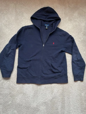 Marinblå hoodie från Polo Ralph Lauren - Snygg marinblå hoodie från Polo Ralph Lauren med dragkedja framtill och klassisk röd logga broderad på bröstet. Hoodien har huva, kängurufickor och är tillverkad i mjukt bomullsmaterial. Perfekt för en avslappnad och stilren look.
