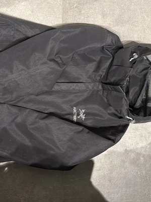 Svart regnjacka från Arc'teryx - Snygg svart regnjacka från Arc'teryx med huva och dragkedja framtill. Jackan har minimalistisk design, justerbar huva och är tillverkad i ett vattentätt material med meshfoder vid halsen. Perfekt för regniga dagar.