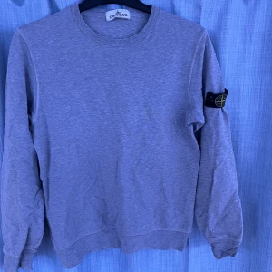 Grå sweatshirt från Stone Island - Säljer en stilren grå sweatshirt från Stone Island med klassisk rund halsringning och ikonisk logotyp-patch på vänster ärm. Tröjan är i mjukt material och har ribbade muddar vid ärmslut och nederkant. Perfekt för en avslappnad look.skriv om ni har några frågor🤝😄