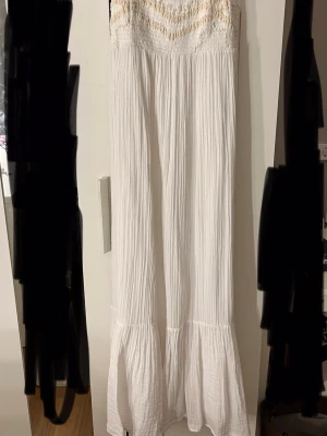 Vit lång sommarklänning med smock - Luftig vit långklänning med smock upptill och volang nertill. Klänningen är ärmlös och har en avslappnad passform i ett mjukt, krinklat tyg. Perfekt för varma dagar och enkel att styla med accessoarer.